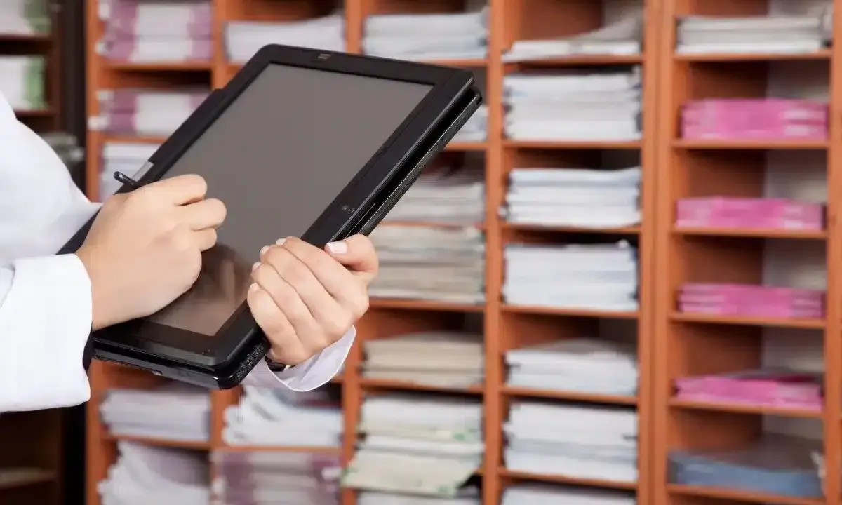 Une personne utilise une tablette pour gérer des documents, illustrant l'archivage électronique, une obligation légale. Les étagères remplies de dossiers rappellent l'importance de la numérisation.