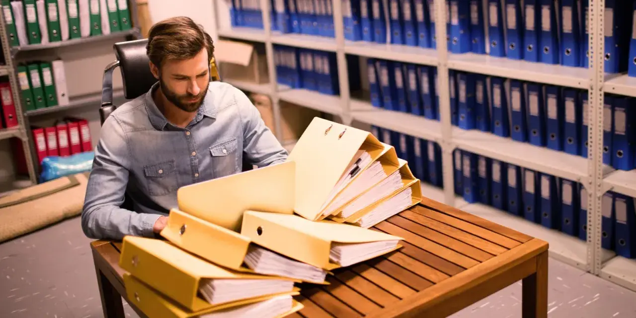 Un homme examine des dossiers dans un **archive papier**. Des étagères remplies de classeurs bleus et verts s'étendent à l'arrière-plan.