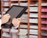 Une personne utilise une tablette pour gérer des documents, illustrant l'archivage électronique, une obligation légale. Les étagères remplies de dossiers rappellent l'importance de la numérisation.