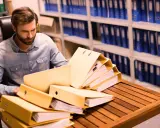 Un homme examine des dossiers dans un **archive papier**. Des étagères remplies de classeurs bleus et verts s'étendent à l'arrière-plan.