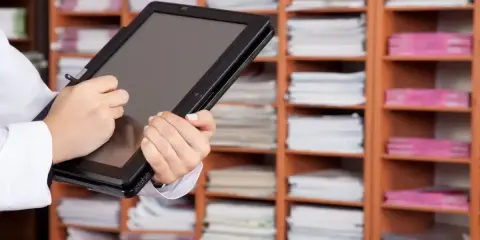Une personne utilise une tablette pour gérer des documents, illustrant l'archivage électronique, une obligation légale. Les étagères remplies de dossiers rappellent l'importance de la numérisation.