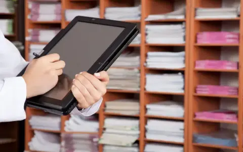 Une personne utilise une tablette pour gérer des documents, illustrant l'archivage électronique, une obligation légale. Les étagères remplies de dossiers rappellent l'importance de la numérisation.