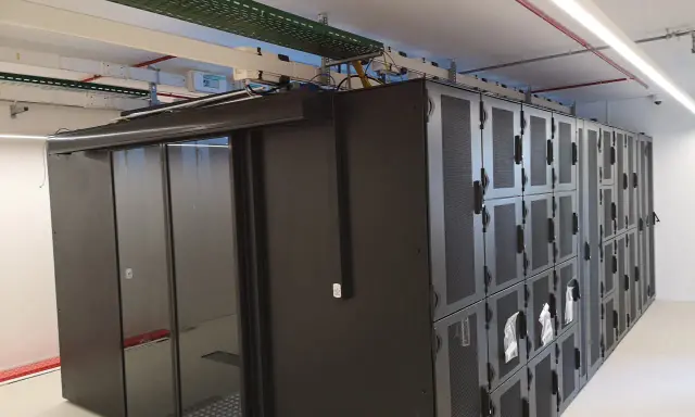 Un micro data center avec des baies de serveurs noires et une porte vitrée. Des câbles et des conduits sont visibles au plafond.
