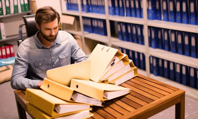 Un homme examine des dossiers dans un **archive papier**. Des étagères remplies de classeurs bleus et verts s'étendent à l'arrière-plan.
