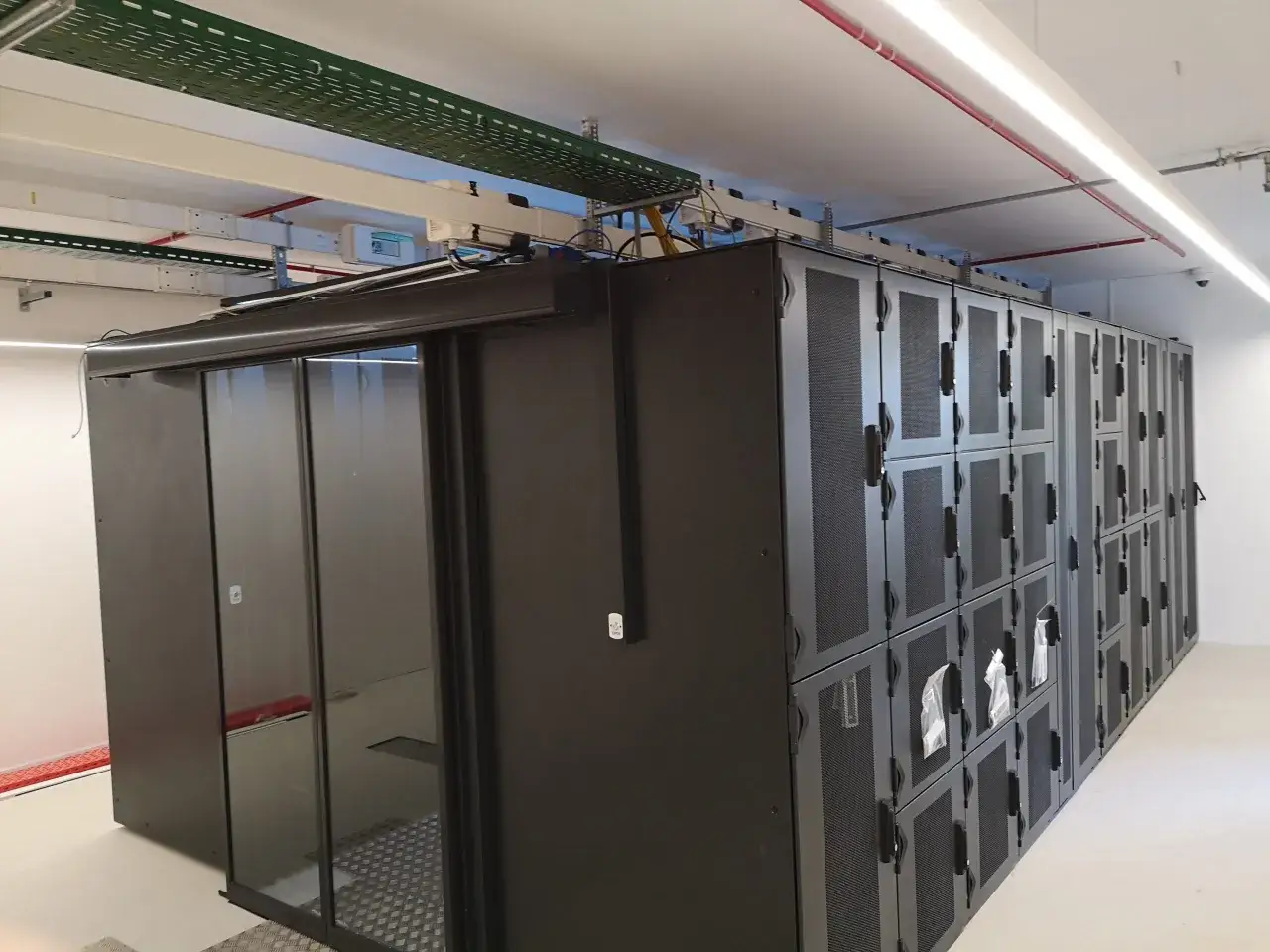 Un micro data center avec des baies informatiques noires et une porte vitrée. Des chemins de câbles sont visibles au plafond.